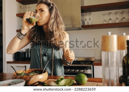 Vegan woman drinking some green juice while having a buddha bowl. Mature woman serving herself a healthy plant-based meal in her kitchen. Senior woman eating clean at home.
