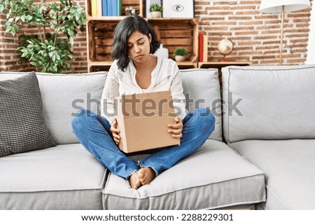 Young hispanic woman sitting on the sofa checking delivery package relaxed with serious expression on face. simple and natural looking at the camera. 