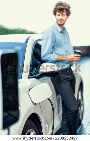 Progressive eco-friendly concept of focus parking EV car at public electric-powered charging station in city with blur background of businessman leaning on recharging-electric vehicle with coffee.