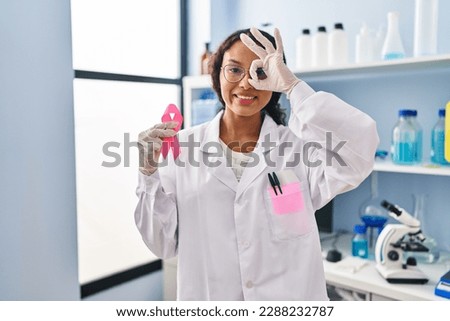 Young hispanic doctor woman working at scientist laboratory holding pink ribbon smiling happy doing ok sign with hand on eye looking through fingers 