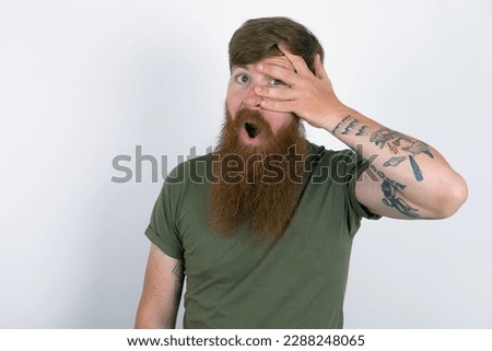 Red haired man wearing green  T-shirt over white studio background peeking in shock covering face and eyes with hand, looking through fingers with embarrassed expression.