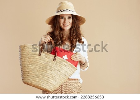Beach vacation. smiling modern 40 years old housewife in white blouse and shorts against beige background with first aid kit, straw bag and summer hat.