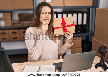 Beautiful woman working at small business ecommerce holding gift serious face thinking about question with hand on chin, thoughtful about confusing idea 
