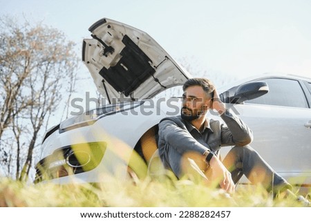 Picture of frustrated man sitting next to broken car with open hood.