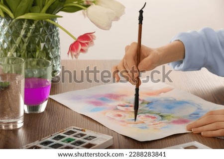 Woman painting flowers with watercolor at wooden table indoors, closeup. Creative artwork