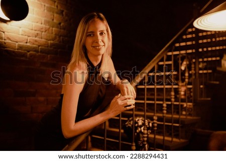 Portrait of young smiling well-groomed slim woman wearing black dress with long fair hair, standing near stairs, leaning on metal bronze railing in restaurant cafe bar, posing. Celebration, party. Royalty-Free Stock Photo #2288294431