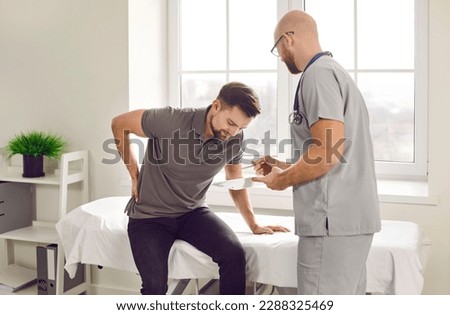 Patient with lower back pain comes to the doctor. Young man suffering from lumbar pain sitting on a medical bed at the clinic and asking a professional physician to help him Royalty-Free Stock Photo #2288325469