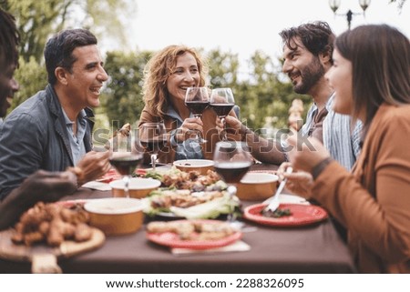 Friends Enjoying Outdoor Dinner - A group of friends aged 25-45 enjoying skewers and wine in the garden. A bearded man and a mature blonde woman toast during a pleasant summer evening. Royalty-Free Stock Photo #2288326095