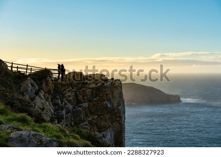 Couple taking a picture on cliffs by the sea at sunset, Asturias, Spain.