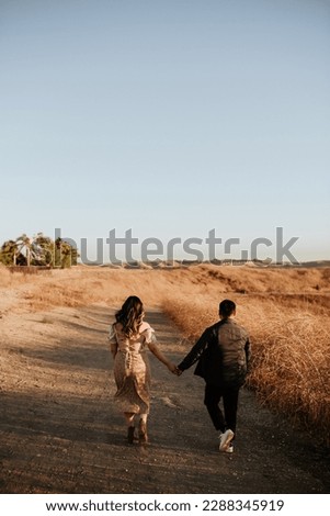 Couple man and woman at their engagement photo session holding hands in a desert like landscape with dried vegetation field at golden hour.