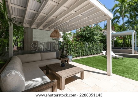 Backyard of a white house located in the Granada neighborhood in Coral Gables, Miami, FL, USA, covered patio with outdoor furniture, short grass area, outdoor kitchen, palms and sun loungers with blue