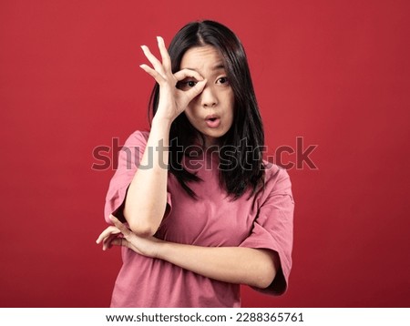 A studio portrait of a young Indonesian (Asian) woman wearing a pink shirt and making the OK sign with her hand in front of her face. Isolated with a red background
