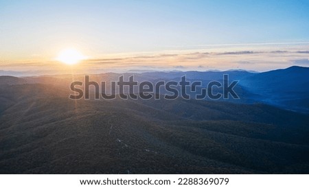 A picturesque sunrise in the mountains. The road winds along the forested mountainside. In the distance a gorge with morning fog rolling in. Shot from a drone.