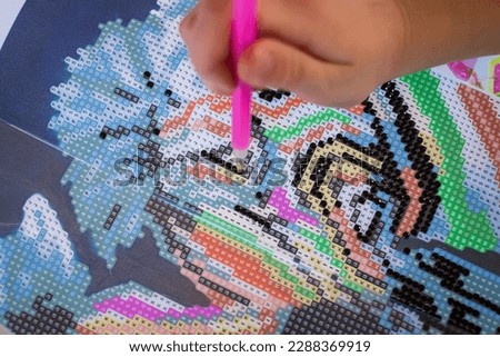 Little girl making diamond painting or pearl mosaic picture. Colorful small pearls stick on image on canvas. Photo series. Trending calming kids leisure activity.
