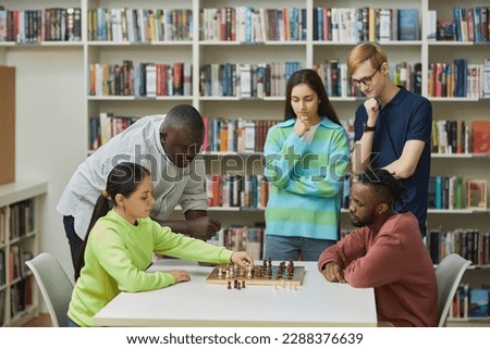 Side view at two young people playing chess in library with diverse group of friends watching