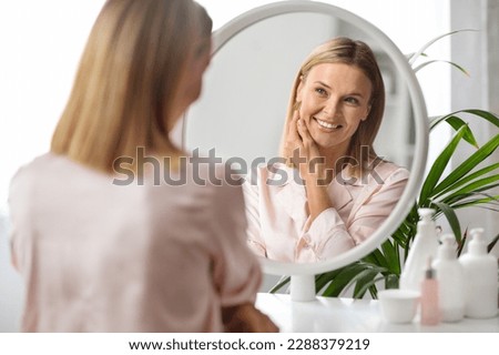 Beauty Care. Attractive Middle Aged Woman Looking In Mirror At Home And Smiling To Her Reflection, Beautiful Mature Blonde Lady Touching Face, Enjoying Flawless Skin After Anti-Aging Treatments Royalty-Free Stock Photo #2288379219