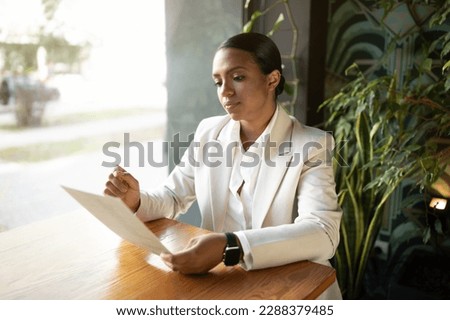 Serious concentrated millennial african american businesswoman in white suit reading document in cafe office with green plants interior. Startup, modern business, work and lifestyle