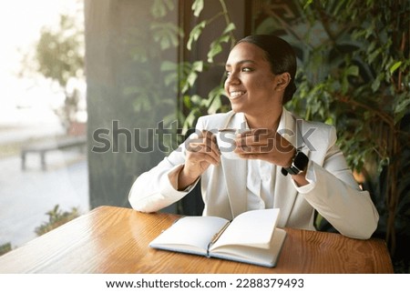 Smiling millennial african american businesswoman in white suit reading book, enjoy cup of coffee in cafe with green plants interior. Break from business, work, lifestyle, rest and relax