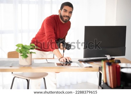 Professional photographer cheerful handsome bearded hispanic young man working on photo shoot at studio, holding dslr camera, using pc computer, smiling at camera, copy space