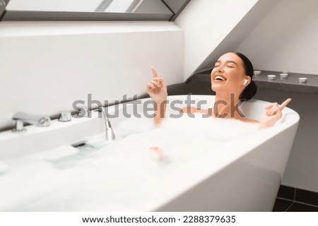 Woman Taking Bath With Foam Enjoying Relaxing Playlist Wearing Earbuds Earphones And Singing Listening To Music Online In Modern Bathroom At Home. Pampering And Relaxation
