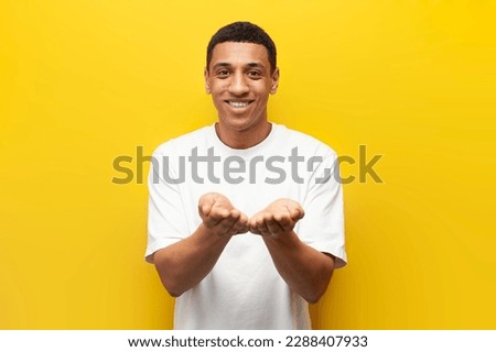 young African American guy holds his hands in front of him on yellow isolated background, man holds nothing and emptiness in his palms
