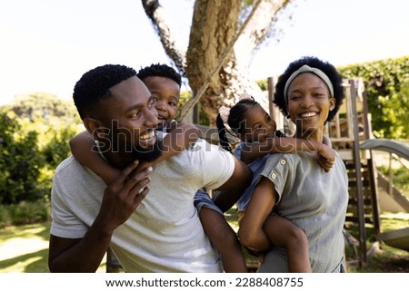 Cheerful african american parents piggybacking son and daughter while standing in playground. Copy space, unaltered, family, togetherness, childhood, enjoyment and weekend concept.