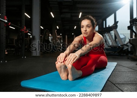 athletic woman in red sportswear sitting on yoga matte in black gym and warming up, girl doing yoga and stretching, attractive woman in fitness club doing flexibility exercise Royalty-Free Stock Photo #2288409897