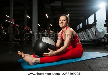 athletic woman in red sportswear sits on yoga matte in black gym and smiles, girl does yoga and warms up, portrait of attractive woman in fitness club Royalty-Free Stock Photo #2288409925
