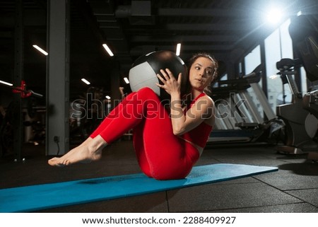 athletic woman in red sportswear pumps abs with medicine ball in black gym, girl does sports on yoga matte, attractive woman in fitness club does exercise with fitness ball Royalty-Free Stock Photo #2288409927