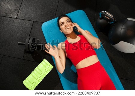 athletic woman in red sportswear lies on yoga matte and talks on the phone in black gym, girl makes phone call in fitness room, attractive woman in fitness club communicates Royalty-Free Stock Photo #2288409937