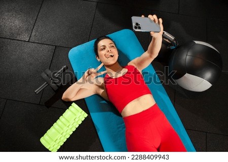 athletic woman in red sportswear lies on yoga matte and takes selfie on smartphone in gym, girl communicates via video call in fitness room, woman in fitness club shows peace gesture on camera Royalty-Free Stock Photo #2288409943