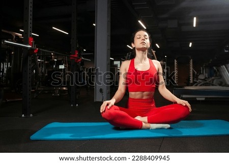 athletic woman in red sportswear sits on yoga matte in black gym and meditates, girl does yoga, attractive woman in fitness club in lotus position Royalty-Free Stock Photo #2288409945