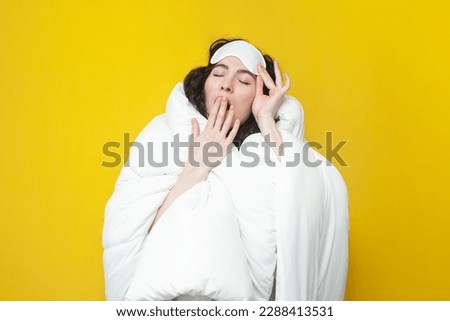 sleepy young girl in sleep mask and with warm soft comfortable blanket yawns on yellow background, tired woman is ready for bed, healthy sleep concept