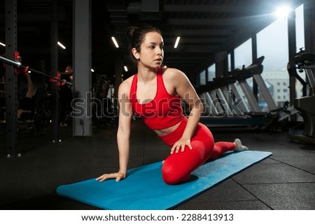 athletic woman in red sportswear sitting on yoga matte in black gym and warming up, girl doing yoga and stretching, attractive woman in fitness club doing flexibility exercise Royalty-Free Stock Photo #2288413913