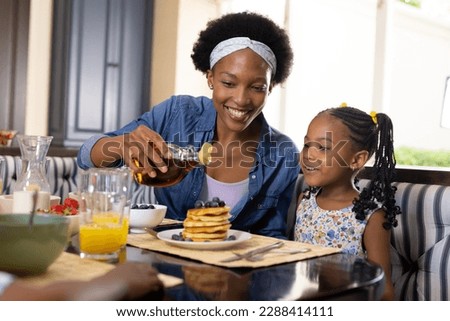 African american smiling mother pouring honey on pancakes while having breakfast with daughter. Unaltered, family, togetherness, childhood, food, fresh, dining table and home concept. Royalty-Free Stock Photo #2288414111