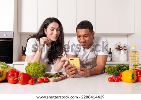 young multiracial couple preparing veggie salad of vegetables and herbs and using smartphone in white kitchen, african american guy with girl choose and order groceries in the phone online Royalty-Free Stock Photo #2288414429
