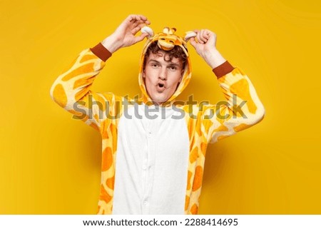 young joyful guy in funny baby giraffe pajamas grimaces and holds his ears on yellow background, man in animal cosplay clothes, pajama party concept