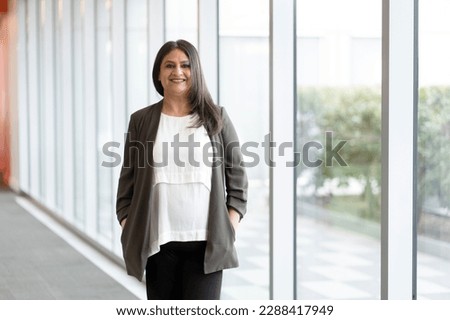 Portrait of confident mature Indian woman standing by large windows in the corporate office, smiling cutely looking at camera. Successful Asian people and business concept Royalty-Free Stock Photo #2288417949