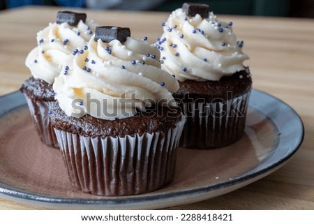 Three chocolate cupcakes with vanilla icing, sprinkles, and chocolate square on a small plate and wooden table. The homemade baked and frosted brownie style cakes swirl in buttercream icing. 