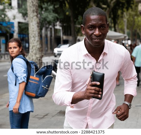 African guy holding stolen wallet running away from young female tourist strolling around city. Street theft concept Royalty-Free Stock Photo #2288421375
