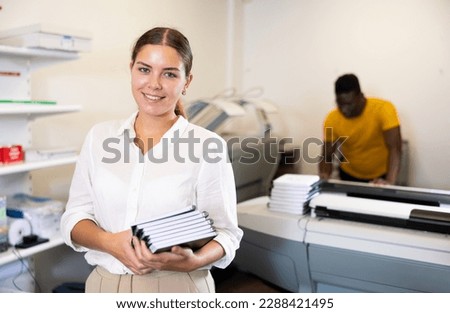 Cheerful young woman in white shirt smiling at the camera and holding new planners in the printing house