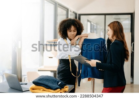 Couple of fashion designers working with fabric and clothing sketches at the studio full of tailoring tools  and holds tablet, notepad and laptop. 
