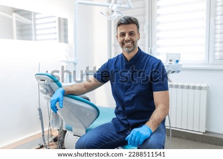 Male dentist posing at clinic over modern cabinet, empty space. Portrait shot of a young smiling dentist sitting in his clinic Royalty-Free Stock Photo #2288511541