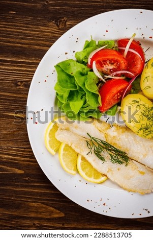 Fish dish - fried cod with boiled potatoes and fresh vegetables on wooden table 