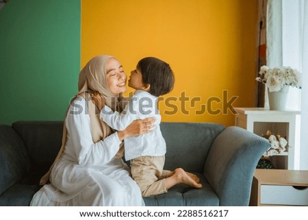 Asian Muslim mom embraces and kiss son sitting on couch in living room