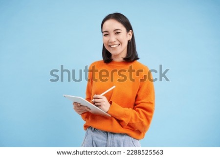 Modern beautiful asian woman with digital tablet and pencil, taking notes, writing on her gadget, doing homework, working, standing over blue background. Royalty-Free Stock Photo #2288525563
