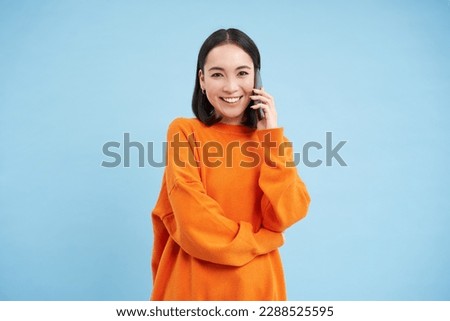 Korean girl in orange sweater talks on mobile phone, speaks on cellphone with happy smiling face, orders delivery, consults with someone on telephone.