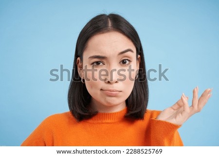 So what. Portrait of korean woman shrugs and raises arm up with skeptical, confused face expression, stands over blue background. Royalty-Free Stock Photo #2288525769