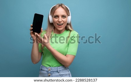photo of a stylish blonde girl in a casual outfit in big white headphones demonstrating a mockup of a smartphone on a blue background