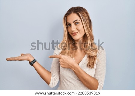 Young blonde woman standing over isolated background amazed and smiling to the camera while presenting with hand and pointing with finger. 